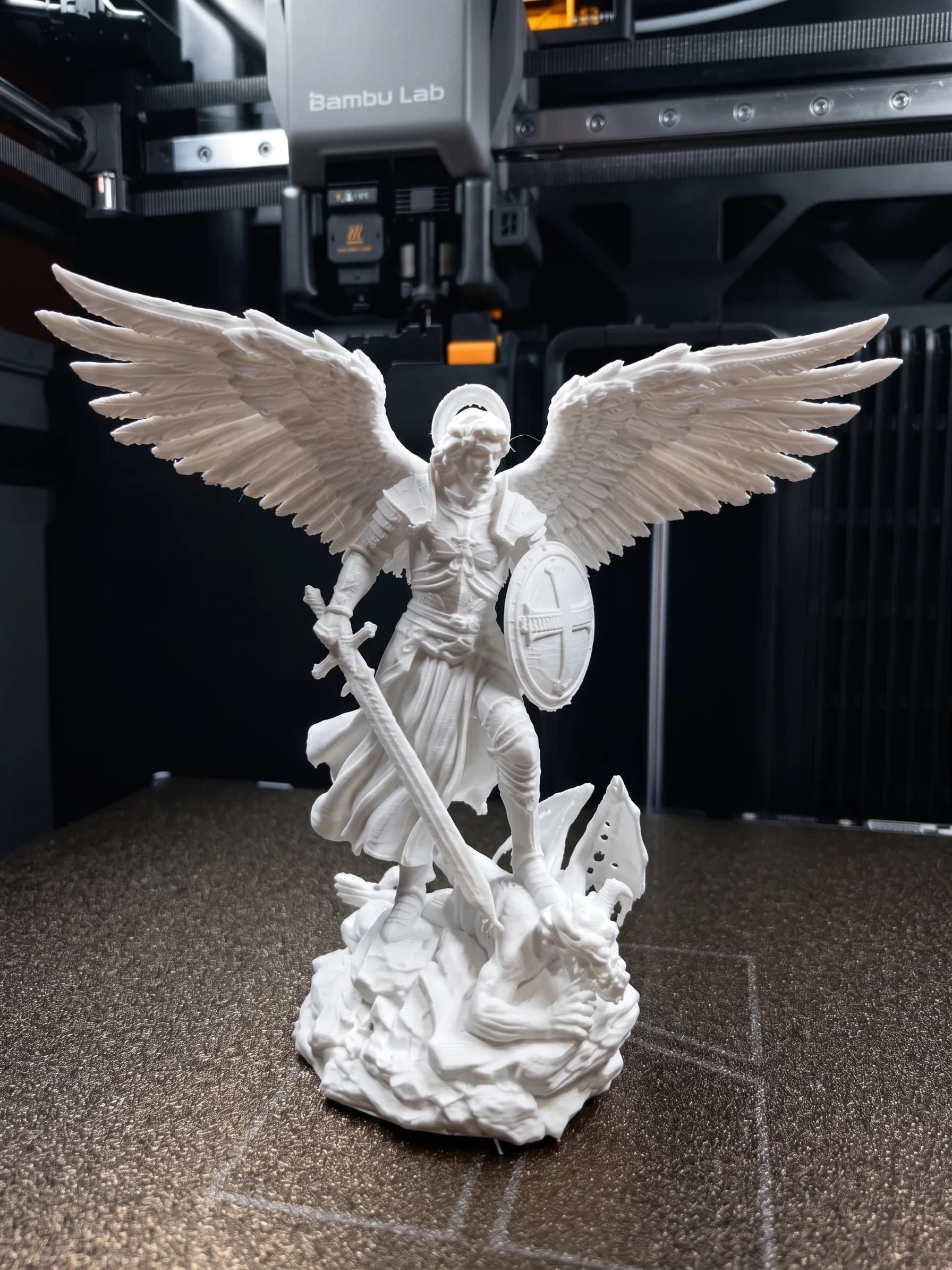 A white archangel figure with wings spread wide, a halo behind the head, a round shield bearing a cross and a sword held down across the body, standing on a rocky base over a fallen dragon